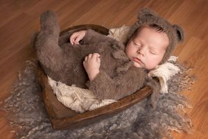 Newborn baby boy in a fuzzy bear outfit laying in a wood heart bowl in Plano, Texas.
