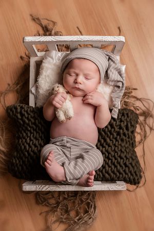 Newborn baby boy laying in a white wood bed wearing sleep pants and hat and holding a small stuffed sheep lovie.