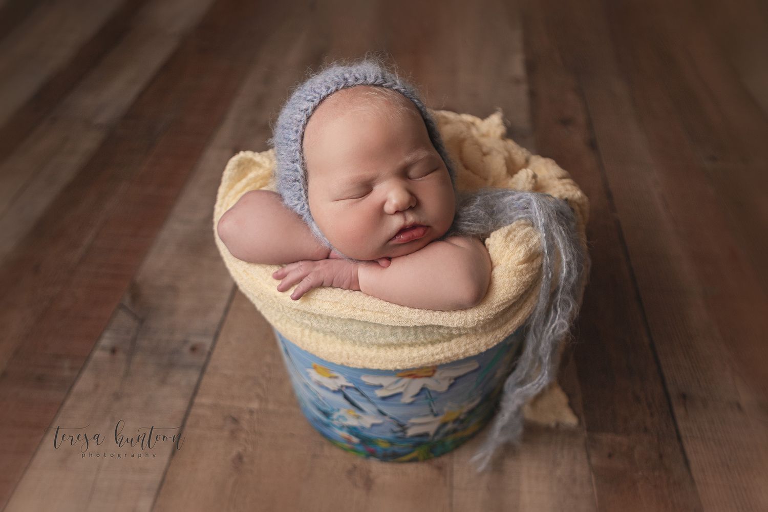 Newborn baby girl with blonde hair in a blue bucket with daisies painted on it wearing a blue bonnet in Dallas, Texas.