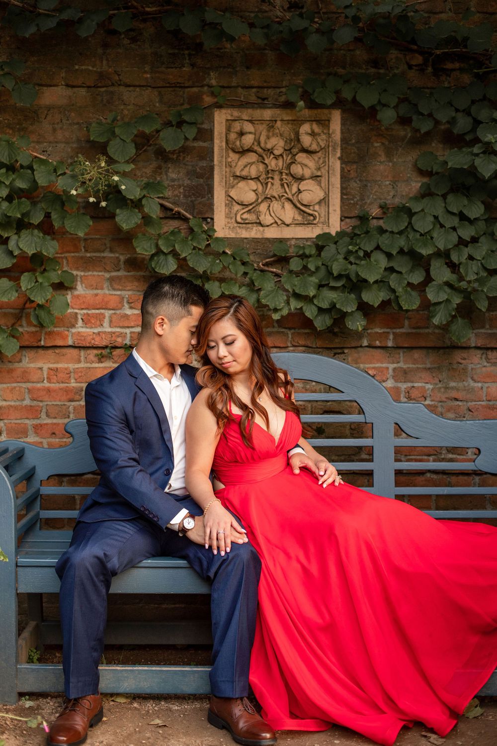 A couple shares an intimate moment on a garden bench against a brick wall with ivy and decorative stone plaque.