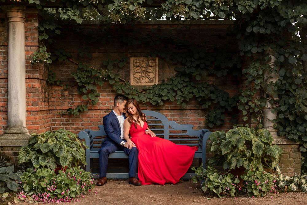 Couple in formal attire sits on blue bench in ivy-covered garden alcove.