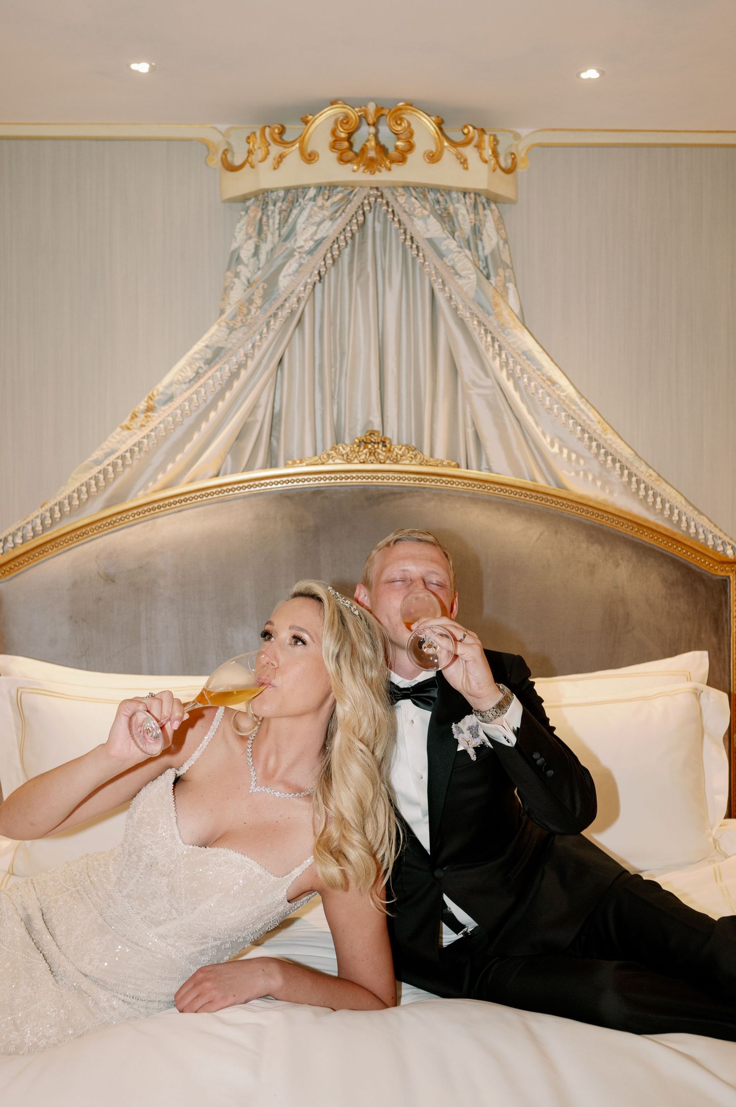 Couple in wedding attire enjoying champagne while relaxing on luxurious bed.