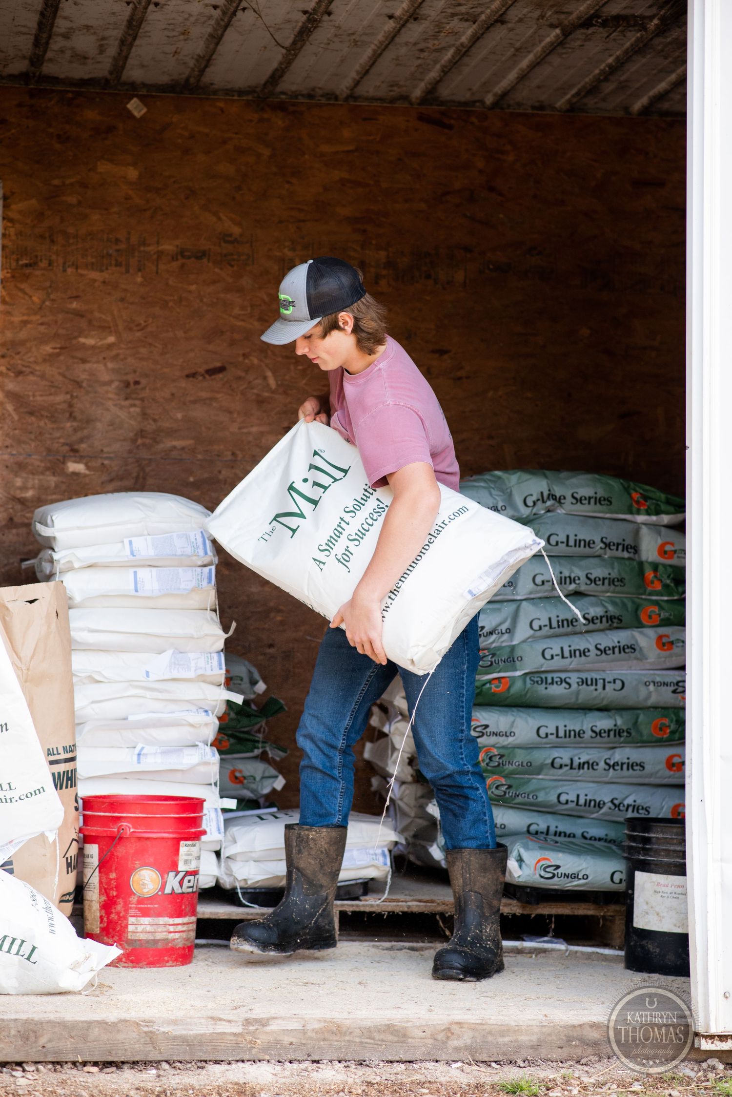 Farm worker carries heavy feed bags in a storage barn while wearing rubber boots and pink shirt.