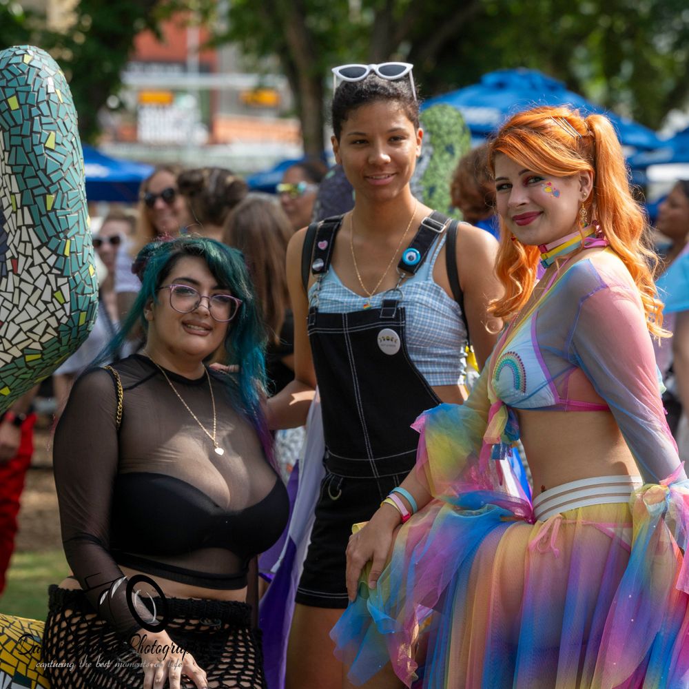 A group in colorful rainbow tulle skirts and festival attire gathers outdoors during a summer celebration.