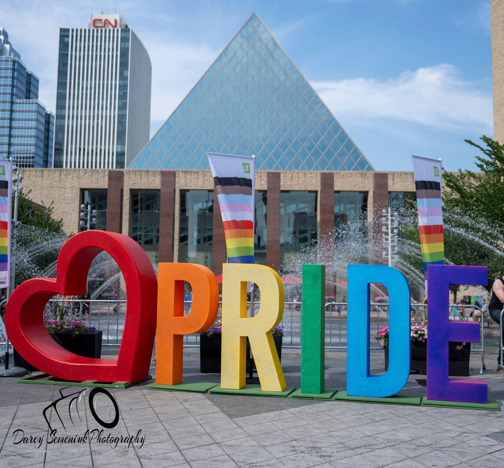 Colorful rainbow Pride letters spelling out 'PRIDE' stand in front of modern glass buildings and pyramids in downtown.