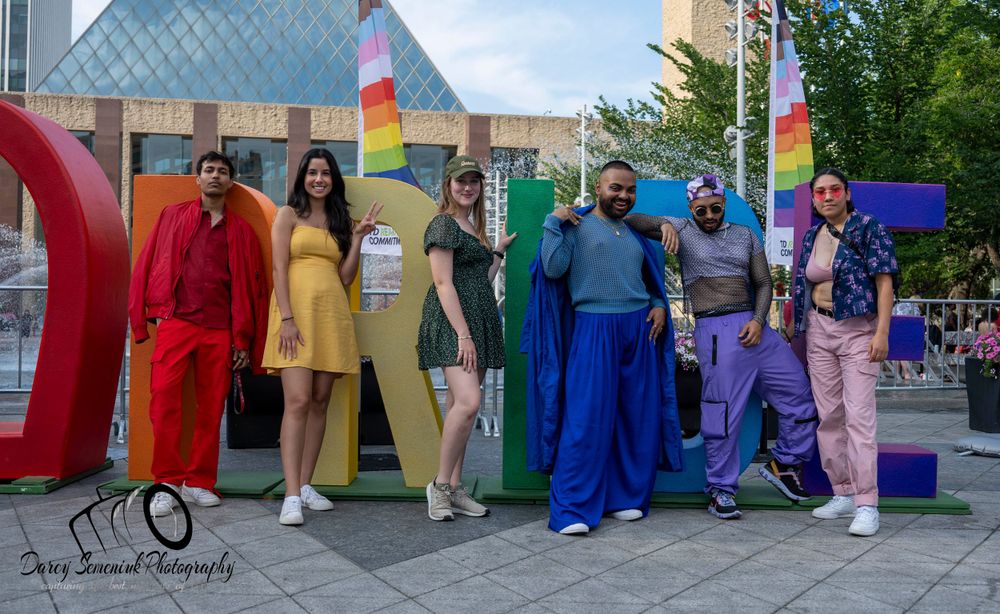 A group stands in colorful outfits arranged like a rainbow spectrum against an outdoor wall.