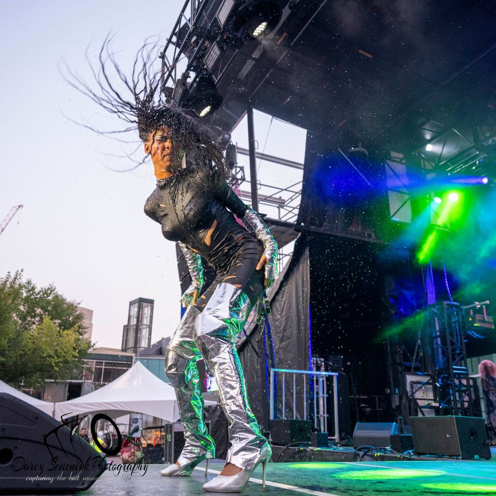 Dynamic performer with long hair jumping on stage with dramatic lighting and green and blue spotlights during live show.