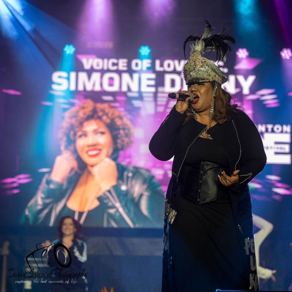 A performer sings on stage with colorful blue and purple lighting while a video screen displays behind them.