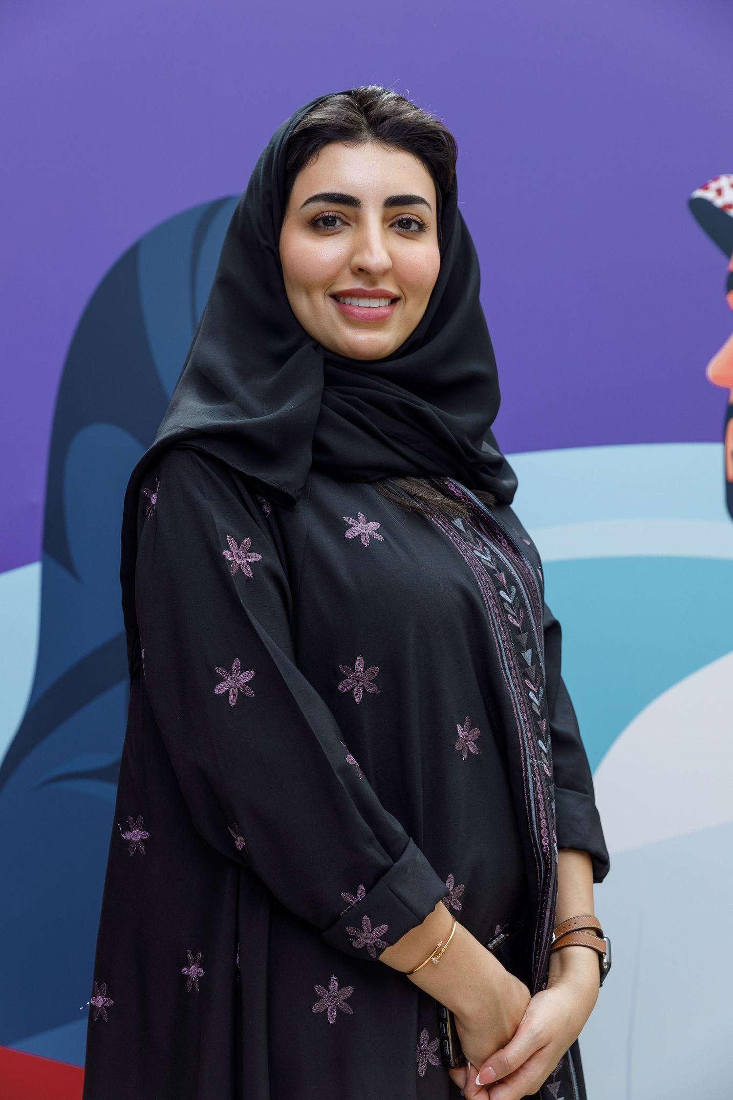 A woman wearing a traditional black abaya with purple embellishments against a colorful backdrop.