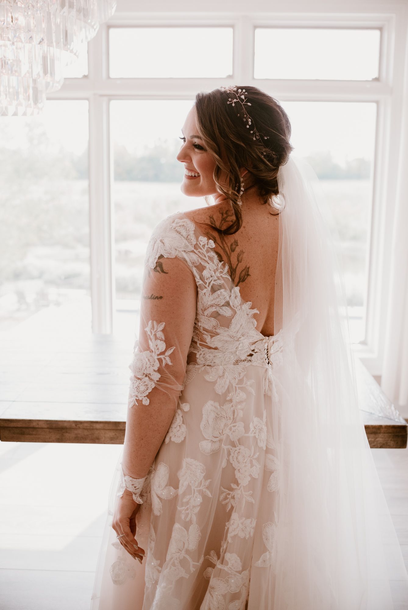 Bride in delicate lace gown standing by bright window with flowing veil.