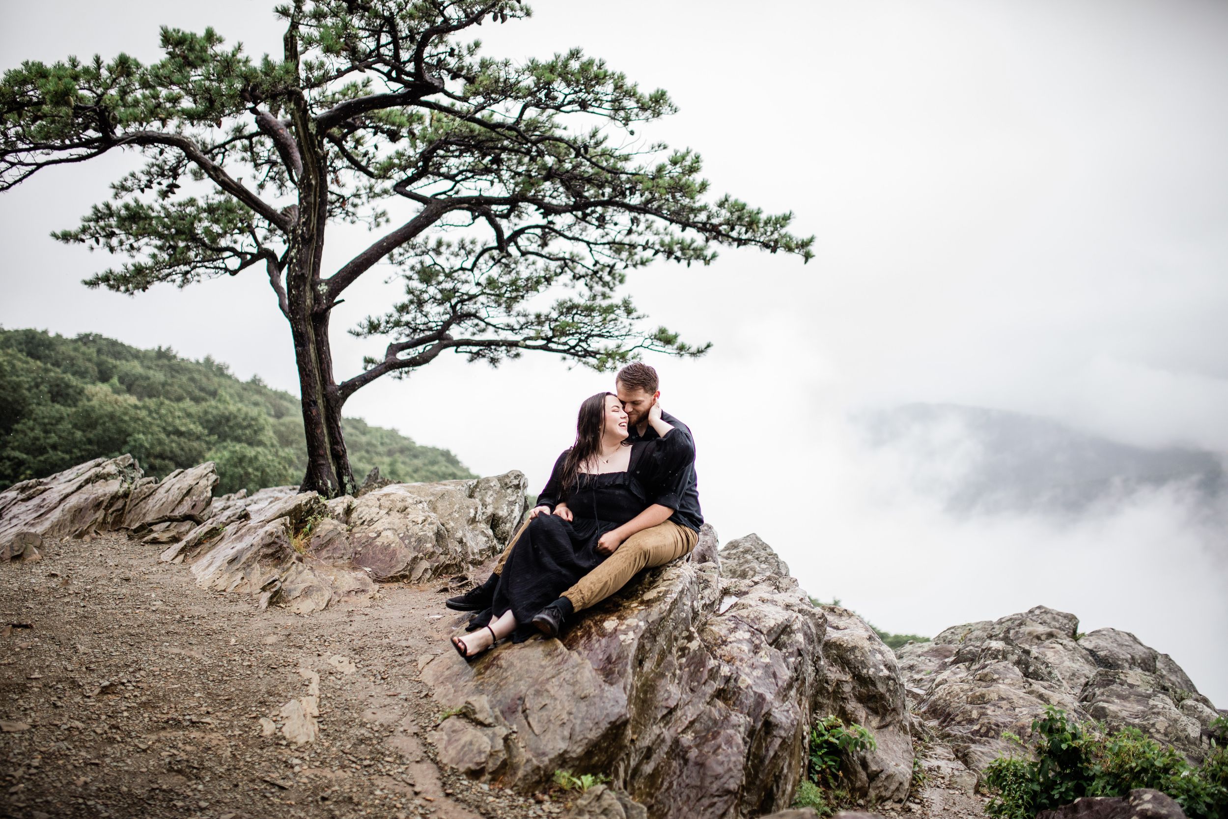 Romantic couple sitting together on rocks beneath pine tree with cloudy mountain landscape.