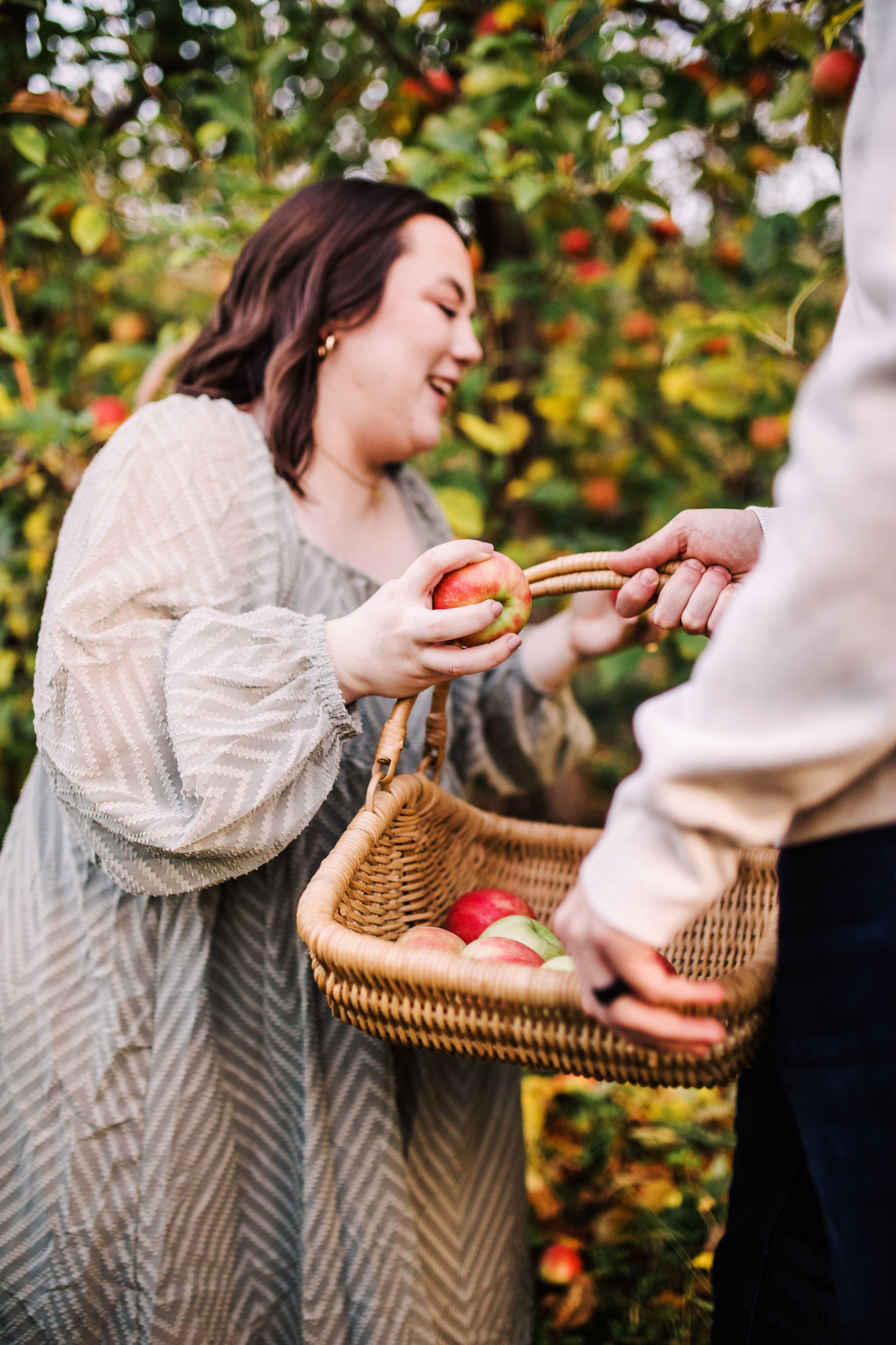 A smiling woman places a red apple into a wicker basket held by her partner in an orchard.
