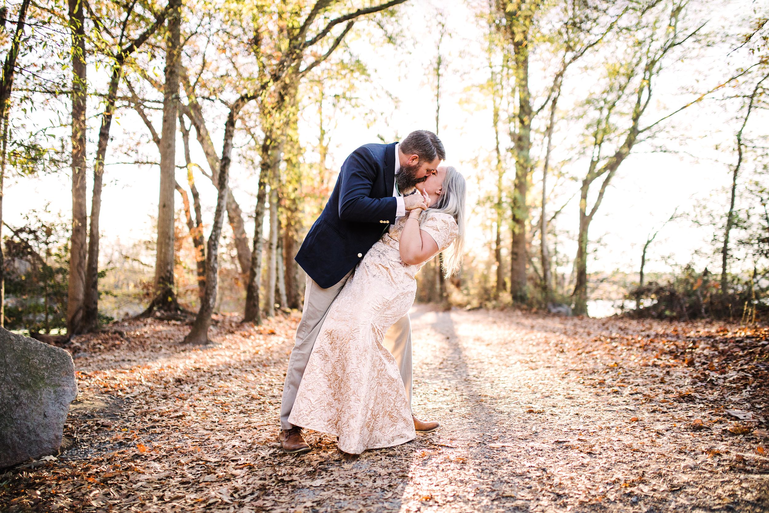 Groom dipping bride for a kiss on a leaf-covered path in a sunlit autumn forest.