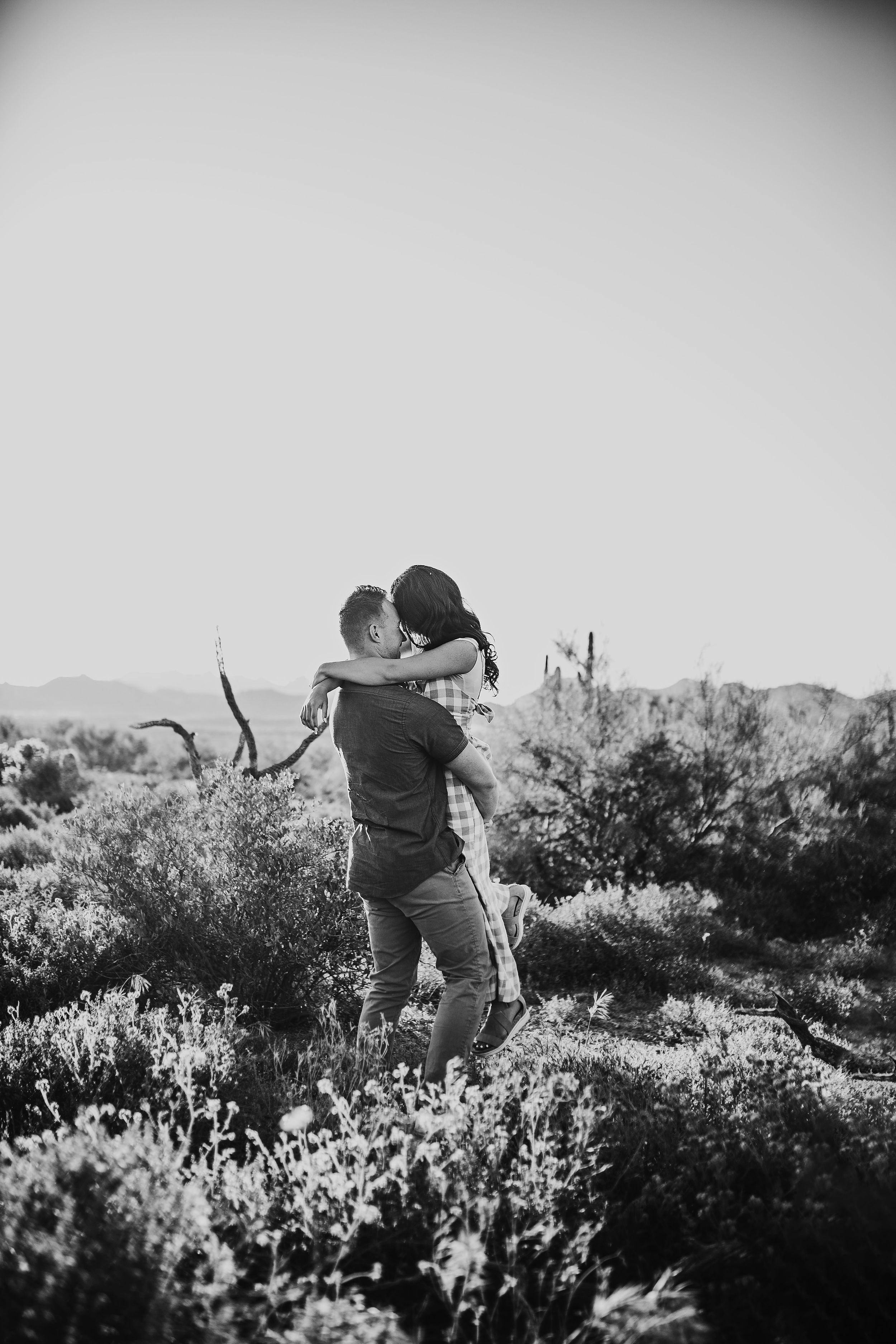Black and white image of man lifting woman in embrace in desert wildflower field.