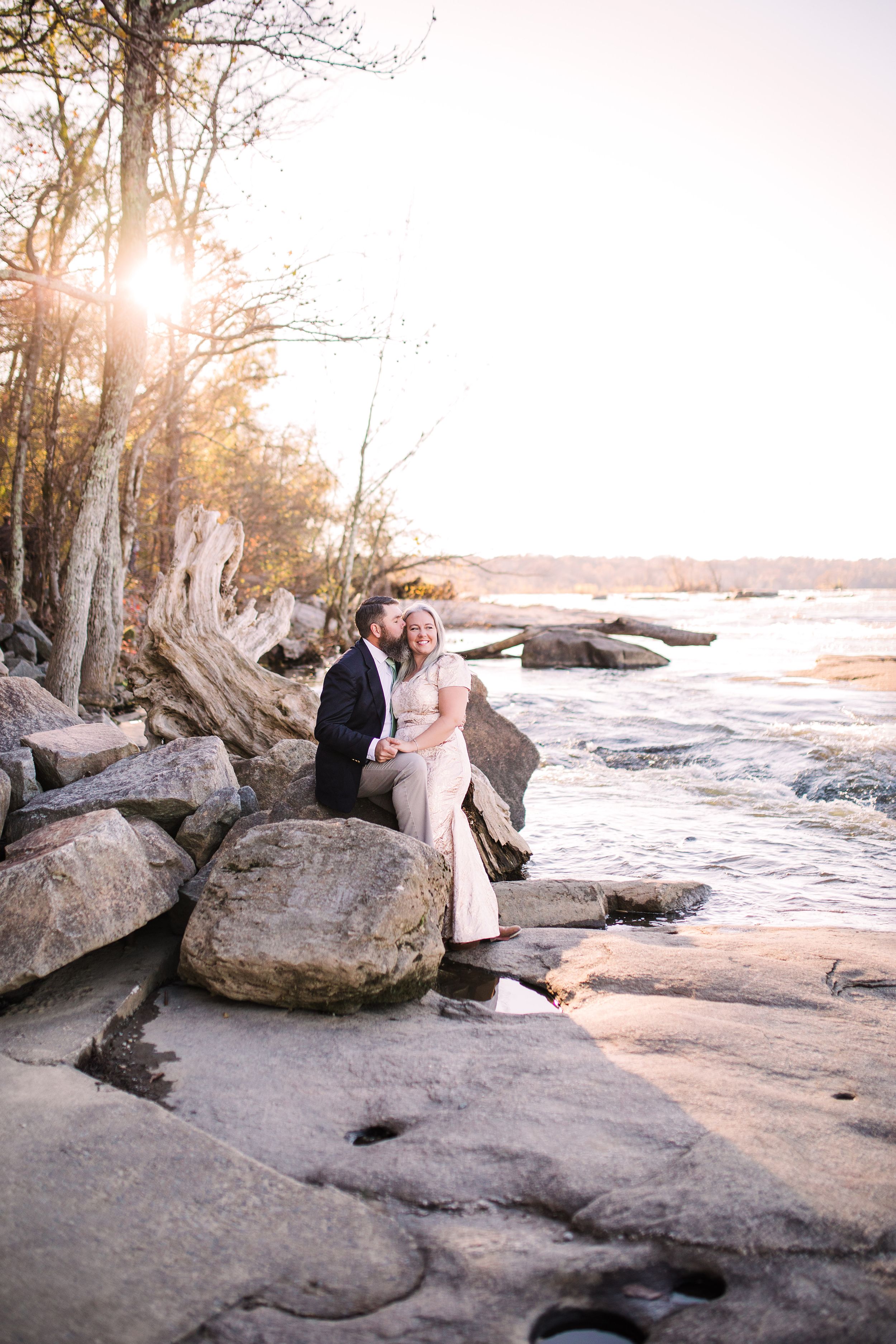 Couple in formal wear sitting on rocks by a river at golden hour sunset.