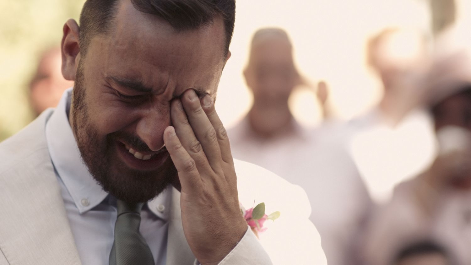Emotional wedding moment captured as groom wipes tears while wearing light colored suit.