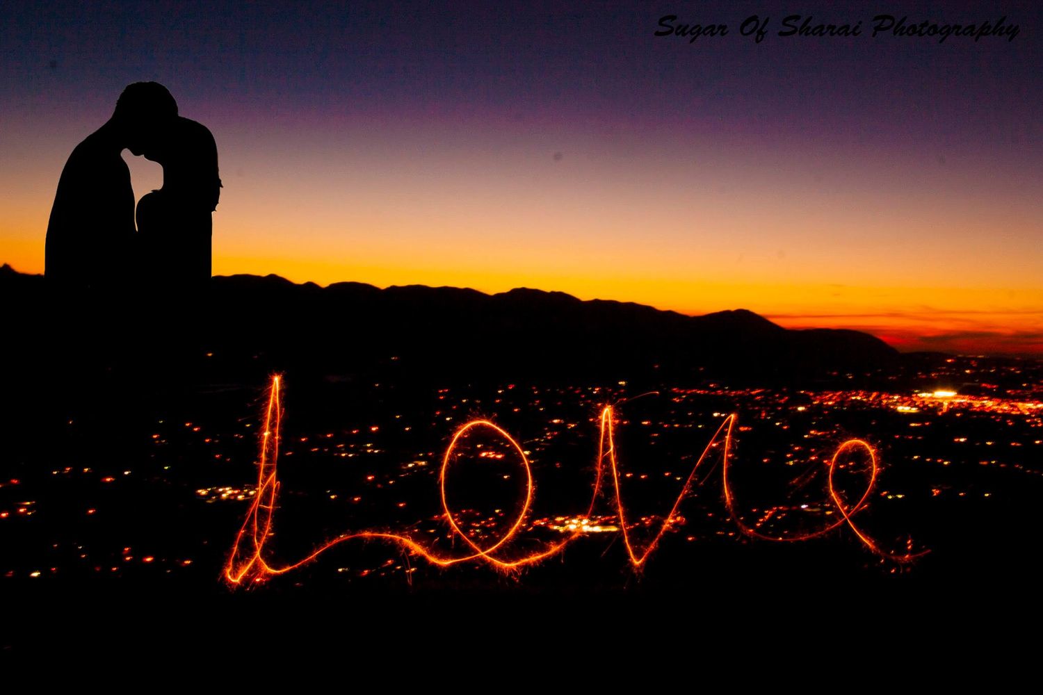 Silhouetted couple against sunset sky with light-painted 'LOVE' text over city lights.