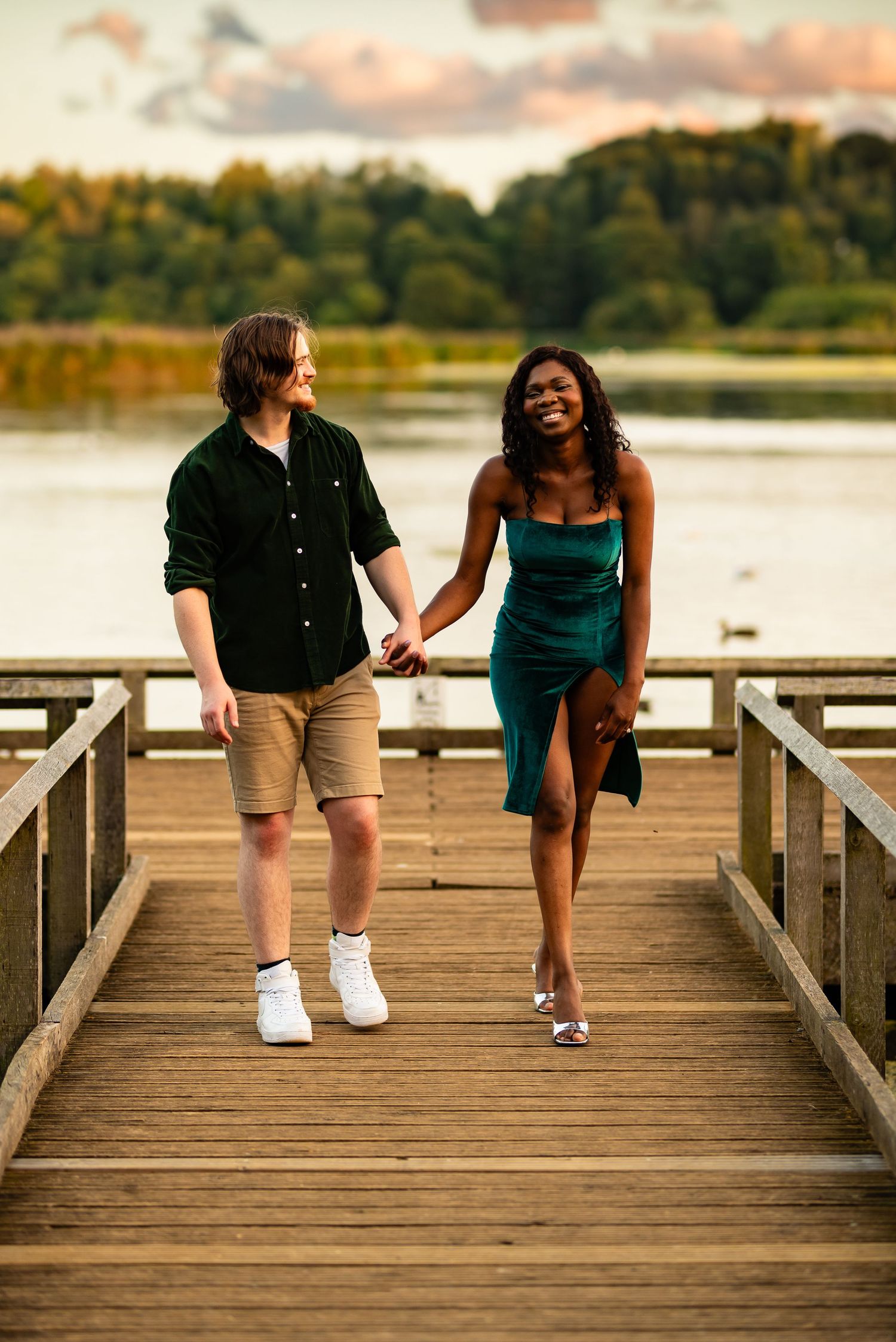 Couple walking hand in hand on wooden pier with lake and autumn foliage in background.
