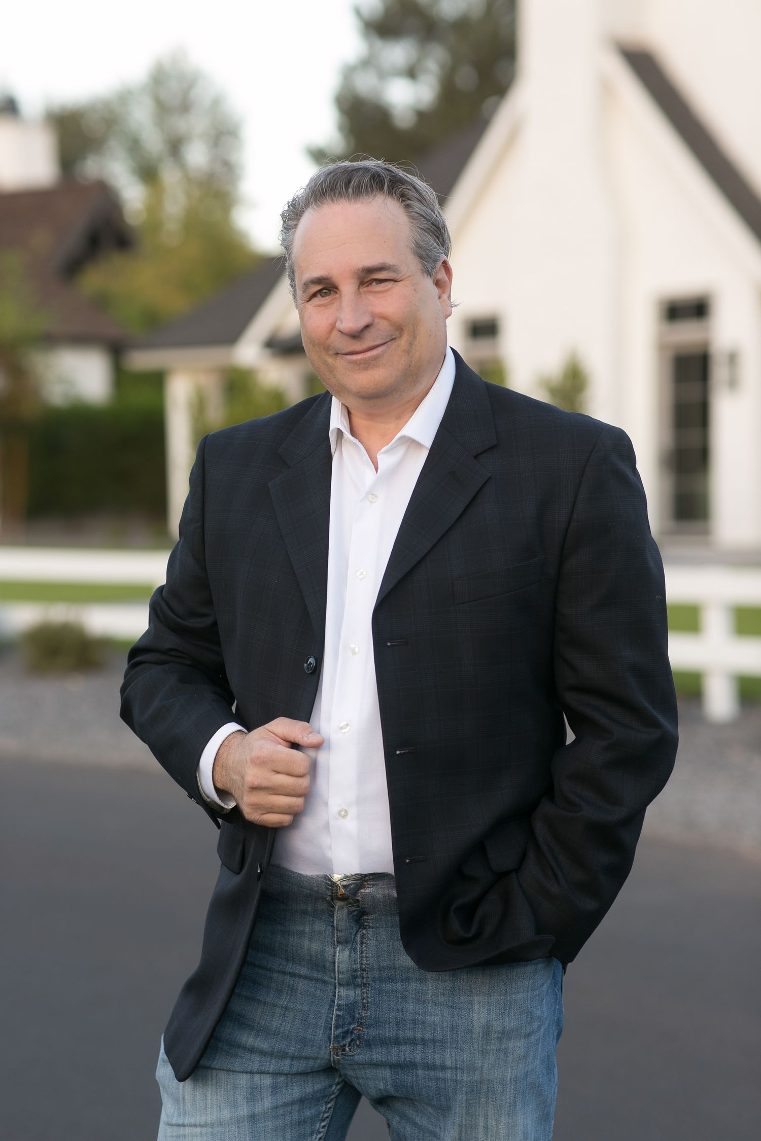 Confident middle-aged man in black blazer and jeans smiling outdoors near a white fence and house.