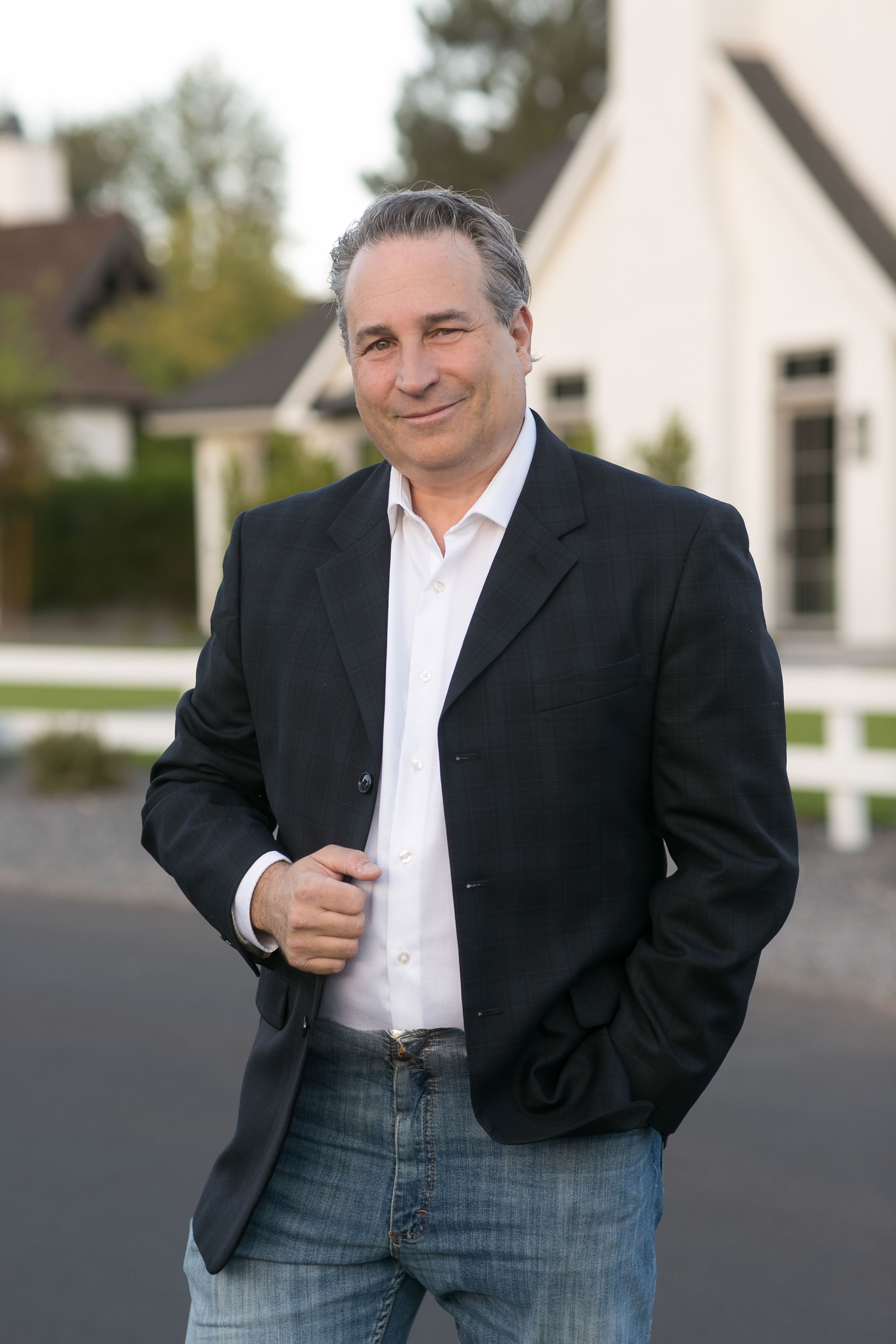 Confident middle-aged man in black blazer and jeans smiling outdoors near a white fence and house.