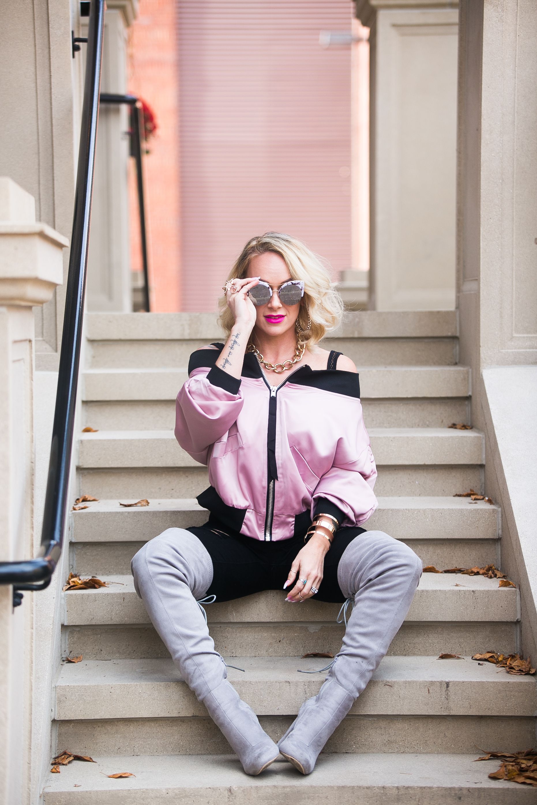 Blonde woman in pink bomber jacket, black shorts, gray over-the-knee boots, sitting on stone steps wearing mirrored sunglasses.