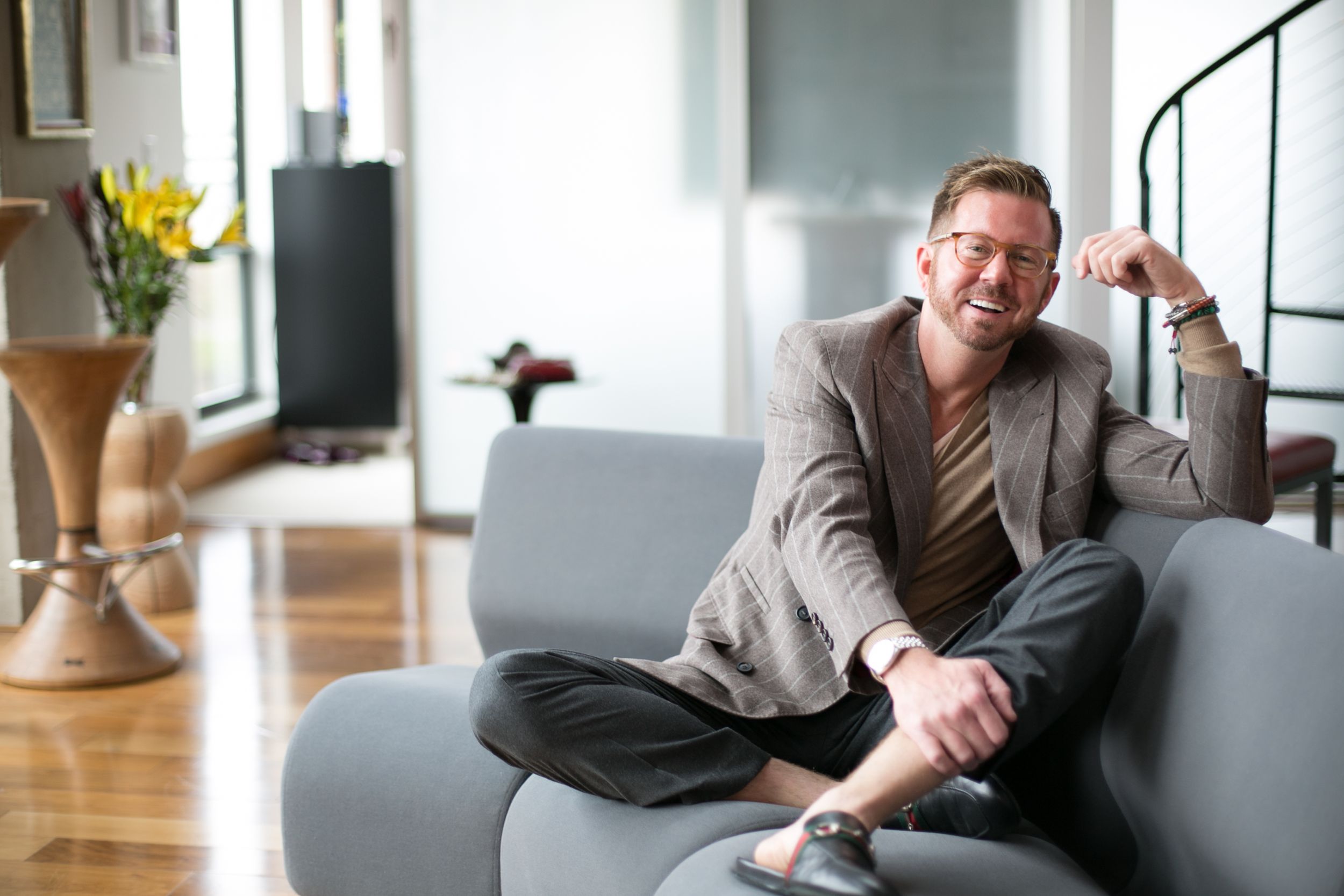 Smiling man in gray blazer sitting cross-legged on modern gray sofa.
