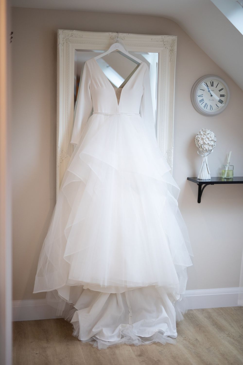 An elegant white wedding dress with long sleeves hangs on display against a beige wall with a clock.