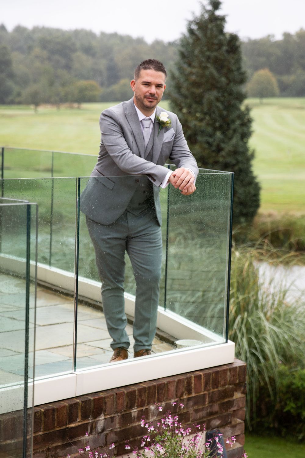 Wedding party member in a grey suit poses by glass railing overlooking a golf course.