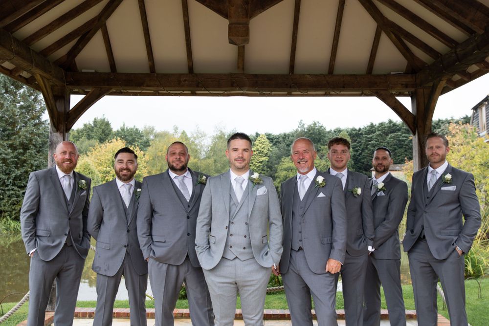 Groomsmen in coordinated grey suits pose under wooden gazebo at outdoor venue.