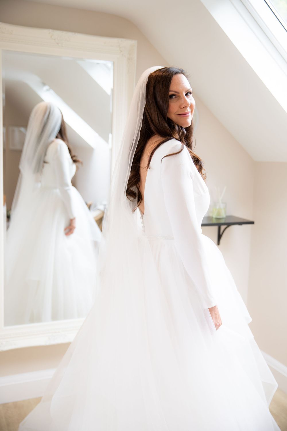 Bride in modern long-sleeved wedding dress posing by mirror in attic room.