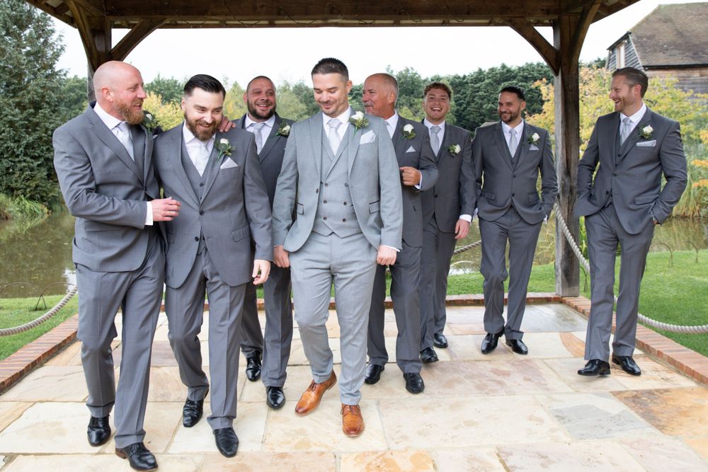 Wedding party in matching grey suits share a candid moment under wooden pavilion.
