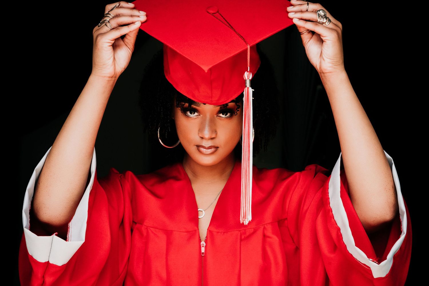 Graduate in bright red cap and gown adjusts mortarboard tassel during graduation photo session.