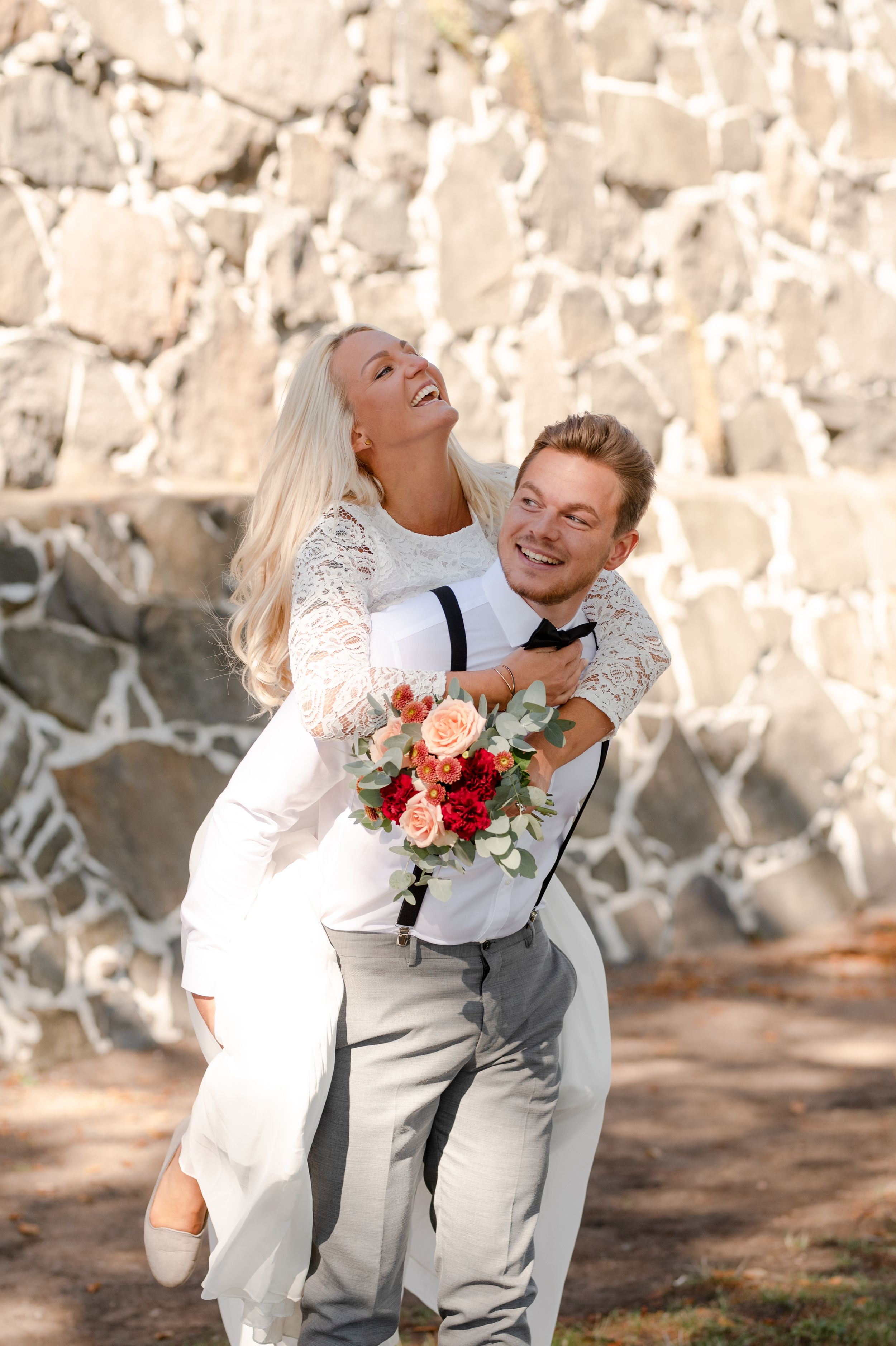 Joyful wedding couple embraces against a stone wall with a vibrant floral bouquet.