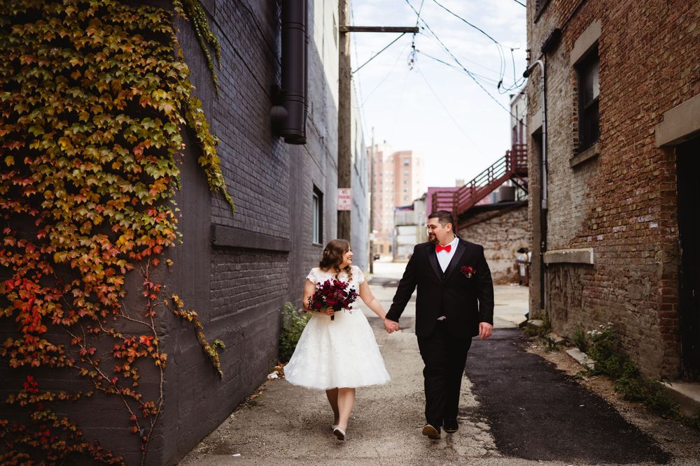 A couple walks hand in hand down an urban alley with fall foliage on brick walls.