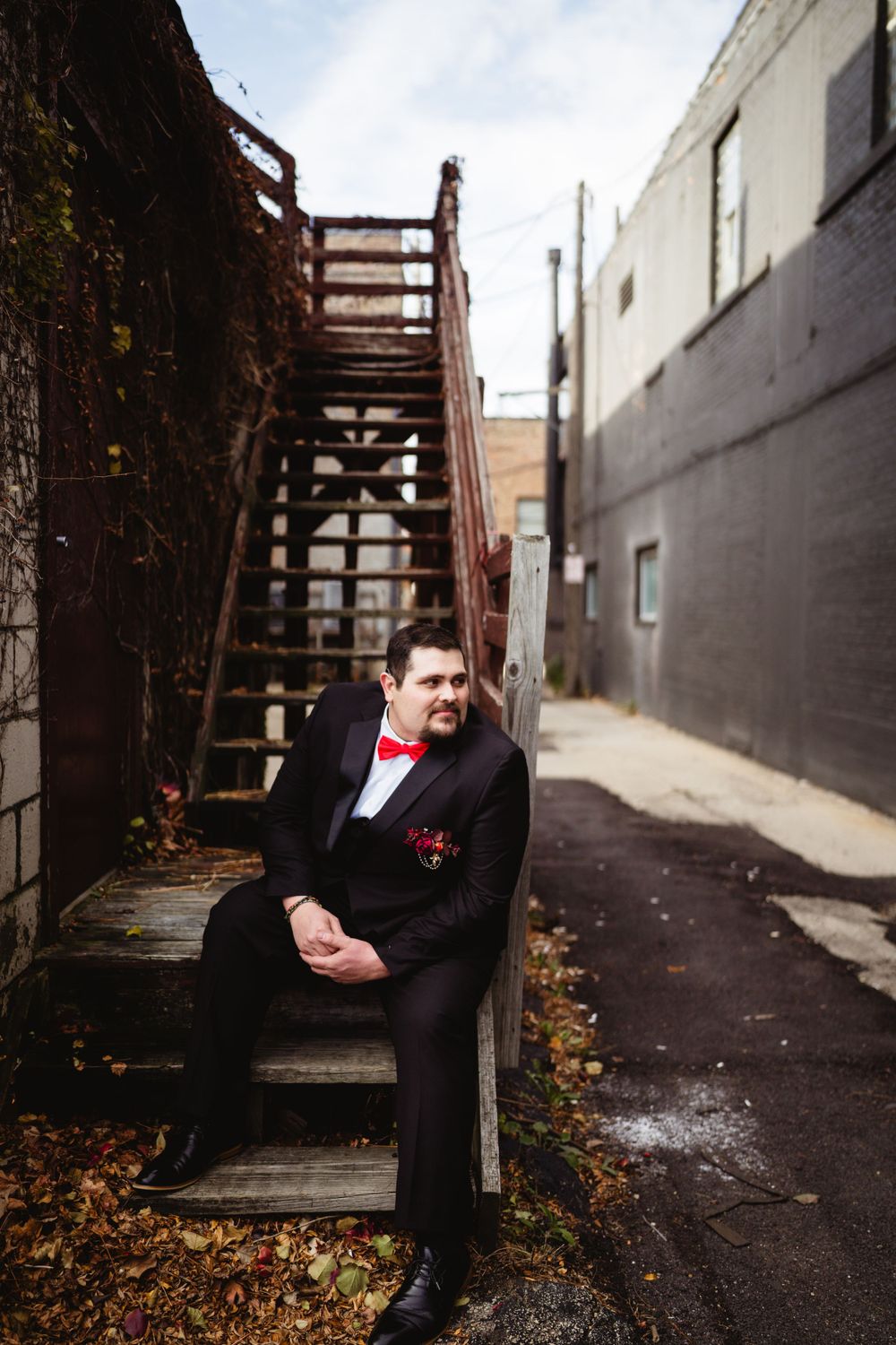 A person in formal attire with red bowtie sits on outdoor wooden stairs in an urban alley.