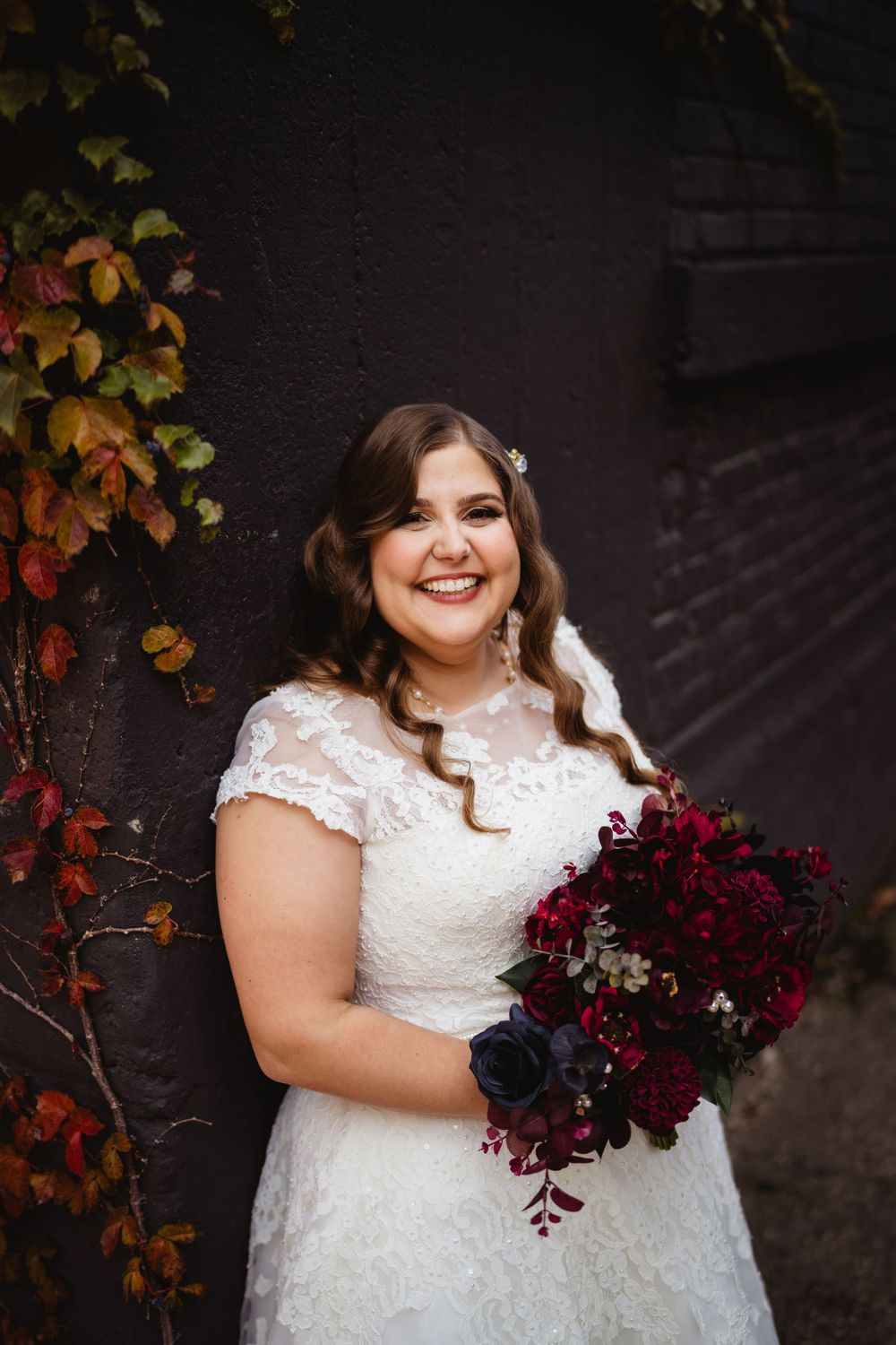 A bride in a lace dress holds a deep burgundy bouquet against an autumn-colored wall.