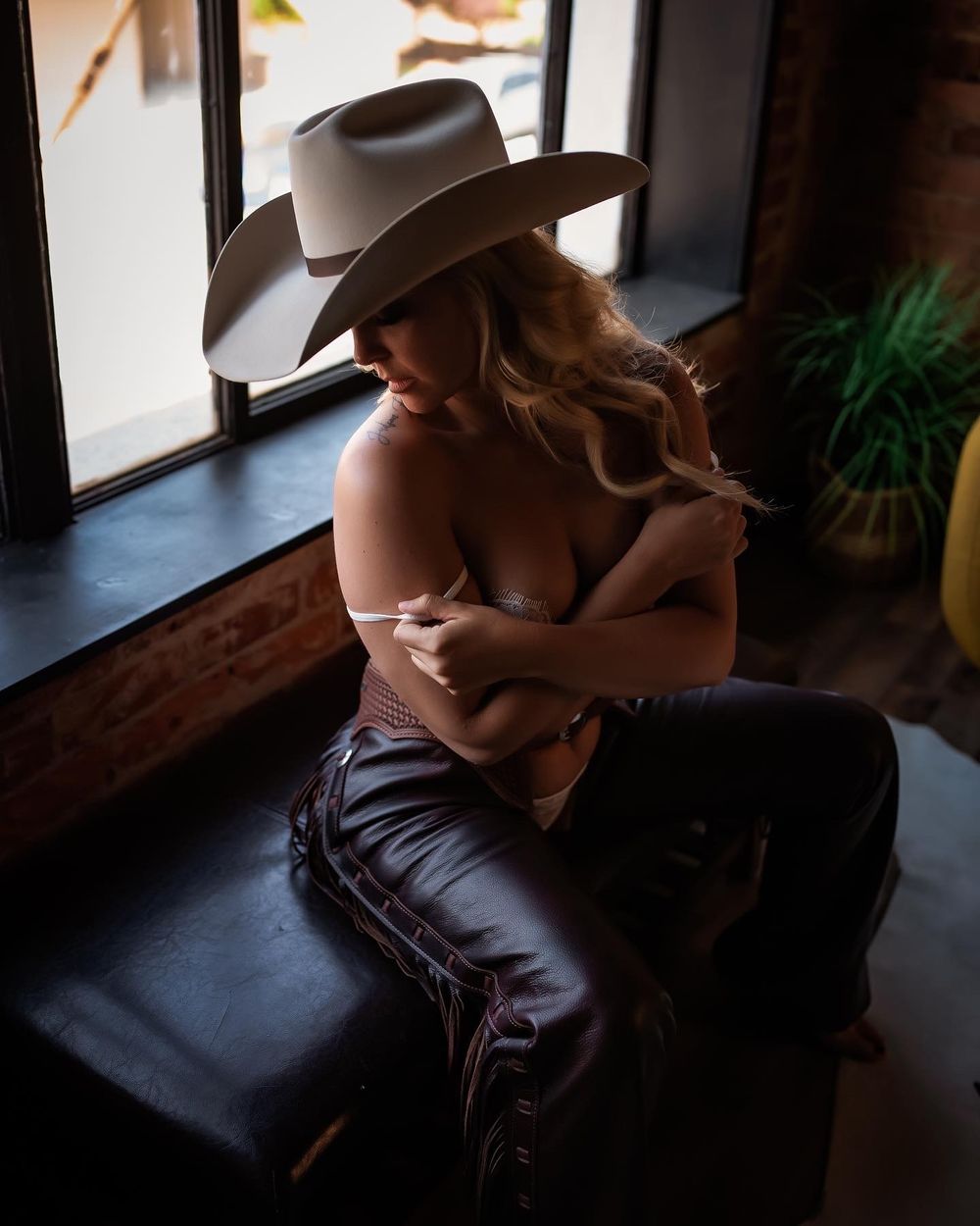 A silhouetted figure in a cowboy hat sits by a window wearing leather pants in moody lighting.