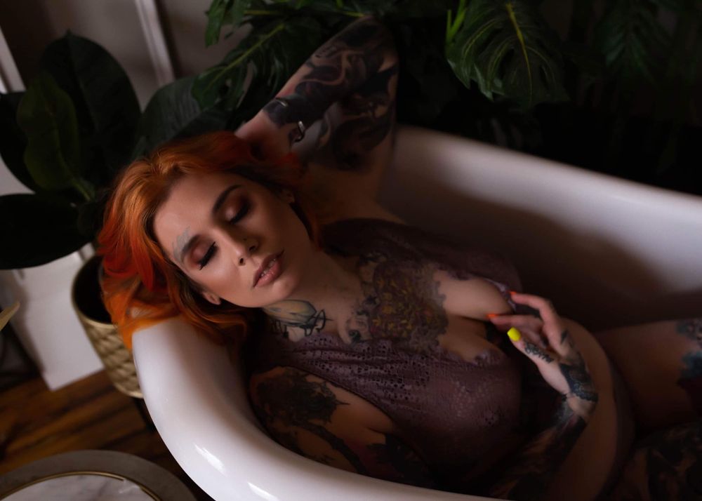 A person relaxes in a white bathtub with reddish hair cascading over the edge in dramatic lighting.