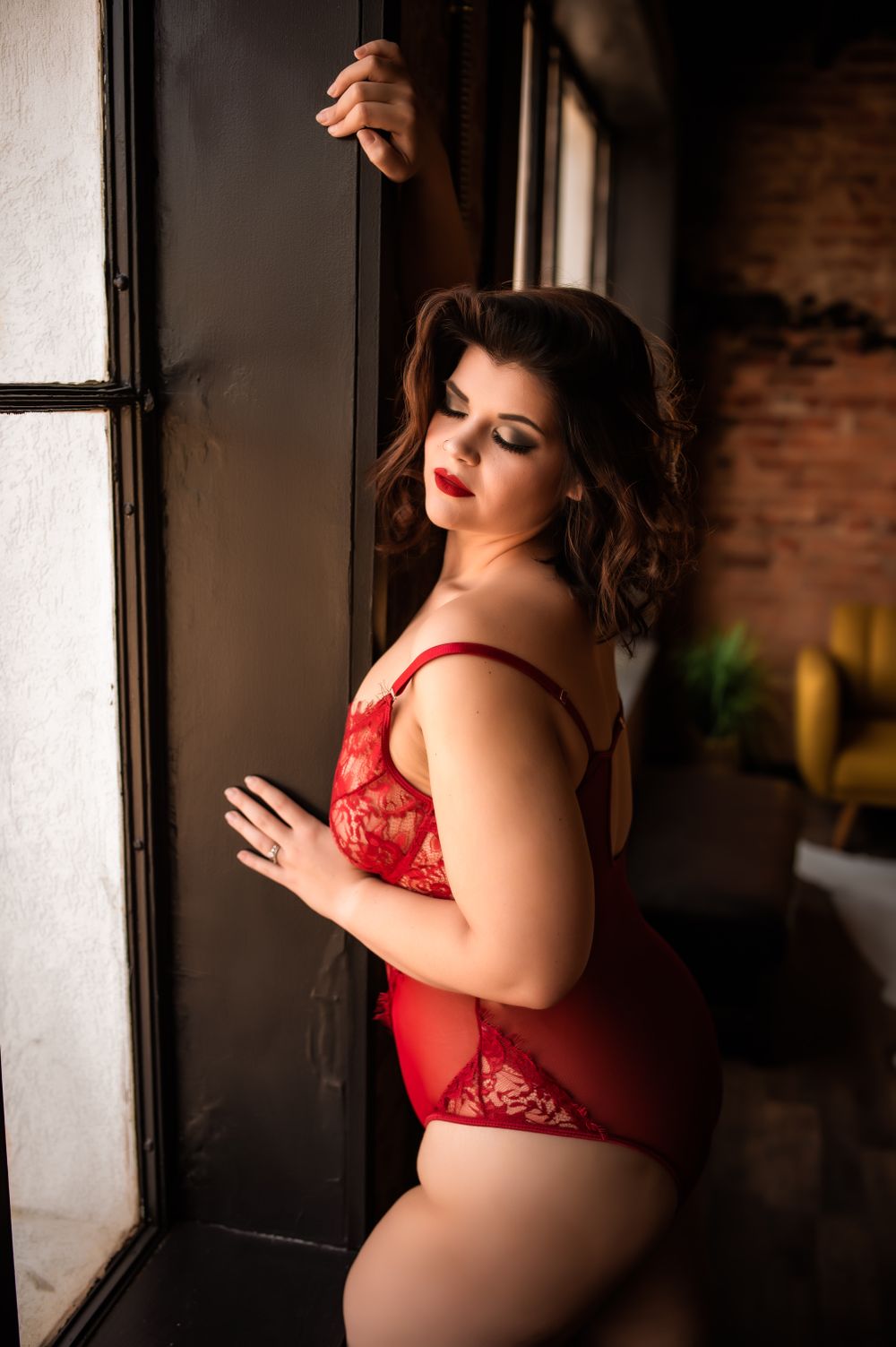 Intimate boudoir photoshoot featuring red lace lingerie in a moody, dimly lit setting with exposed brick walls.