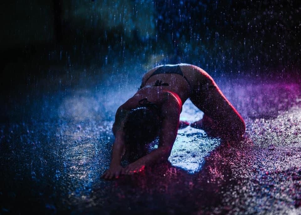 Silhouetted figure in backbend pose illuminated by colorful lights and water spray creating a dramatic dance effect.