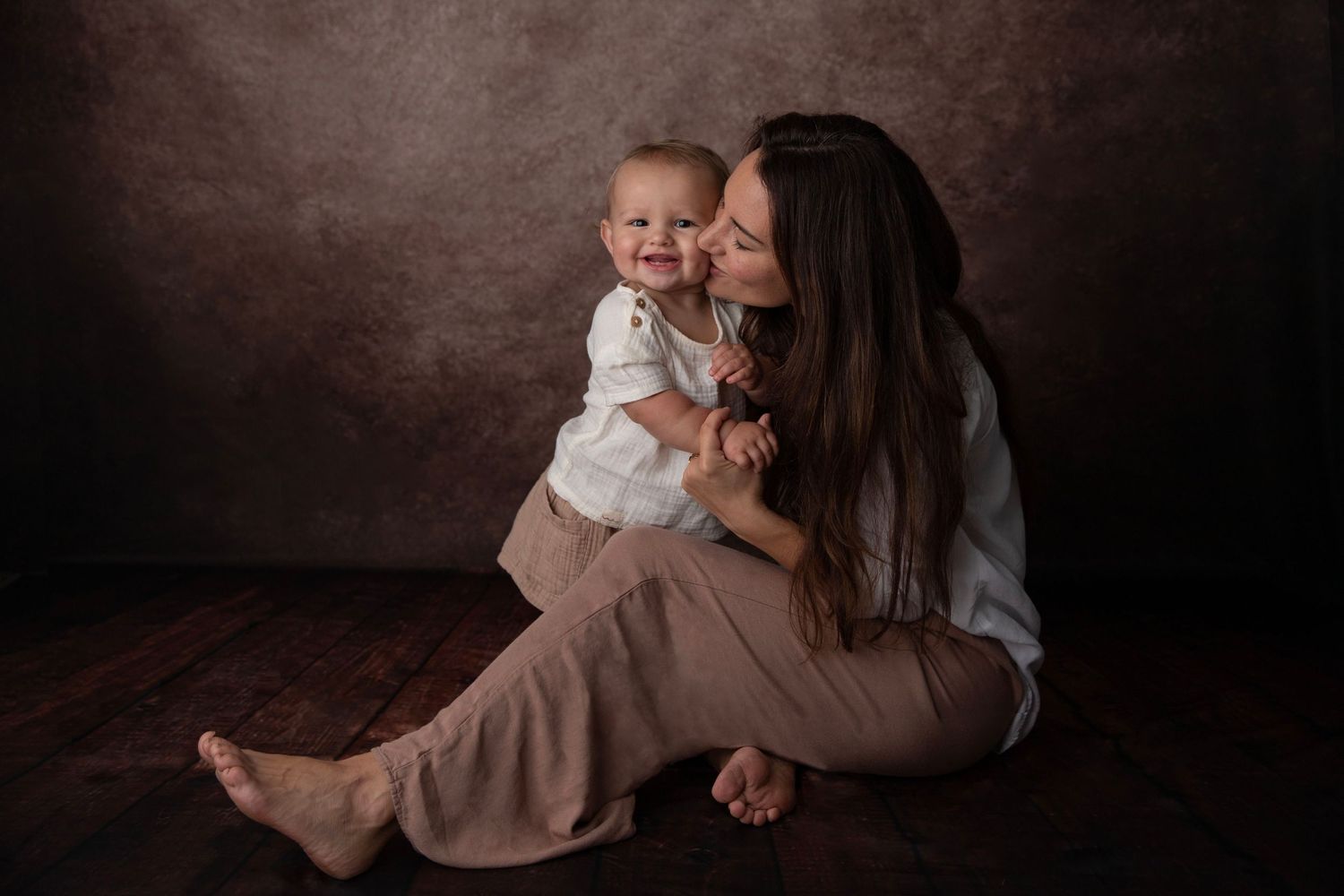 Loving mother sitting on floor kissing smiling baby boy in a professional studio portrait.
