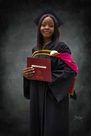 A graduate in academic regalia holds a red diploma cover against a dark background in a formal portrait.