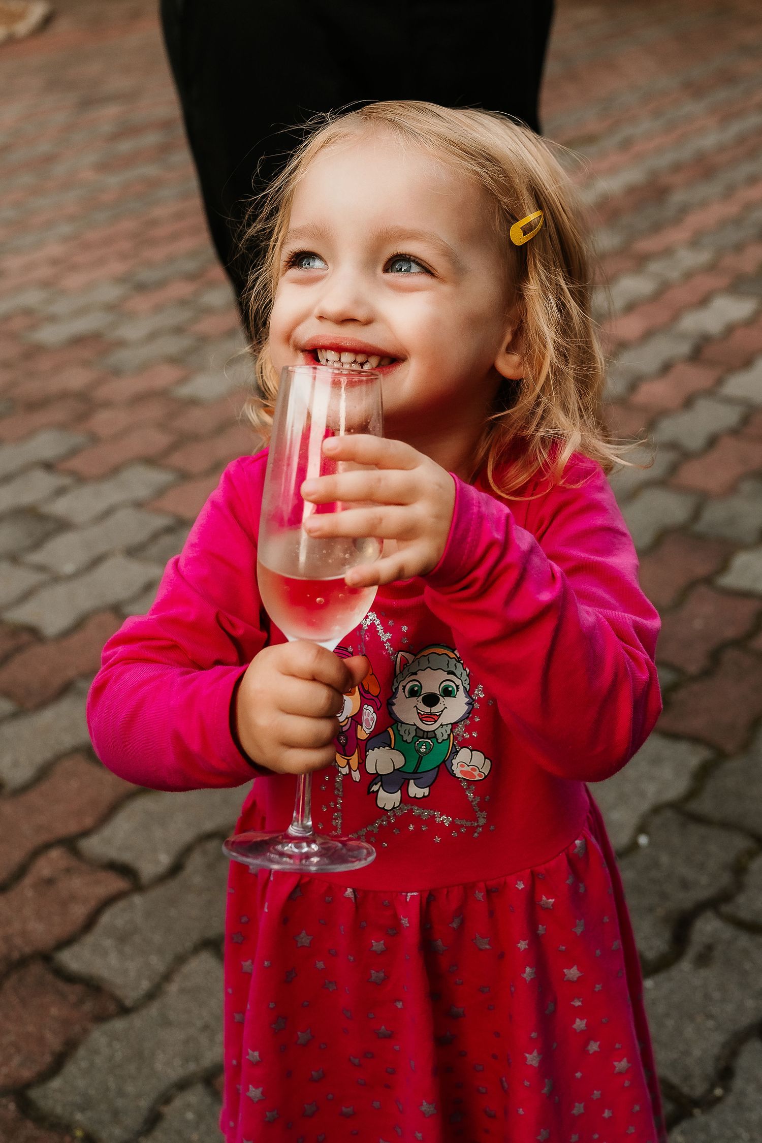 Small child in red dress holding champagne flute filled with pink beverage against brick background.