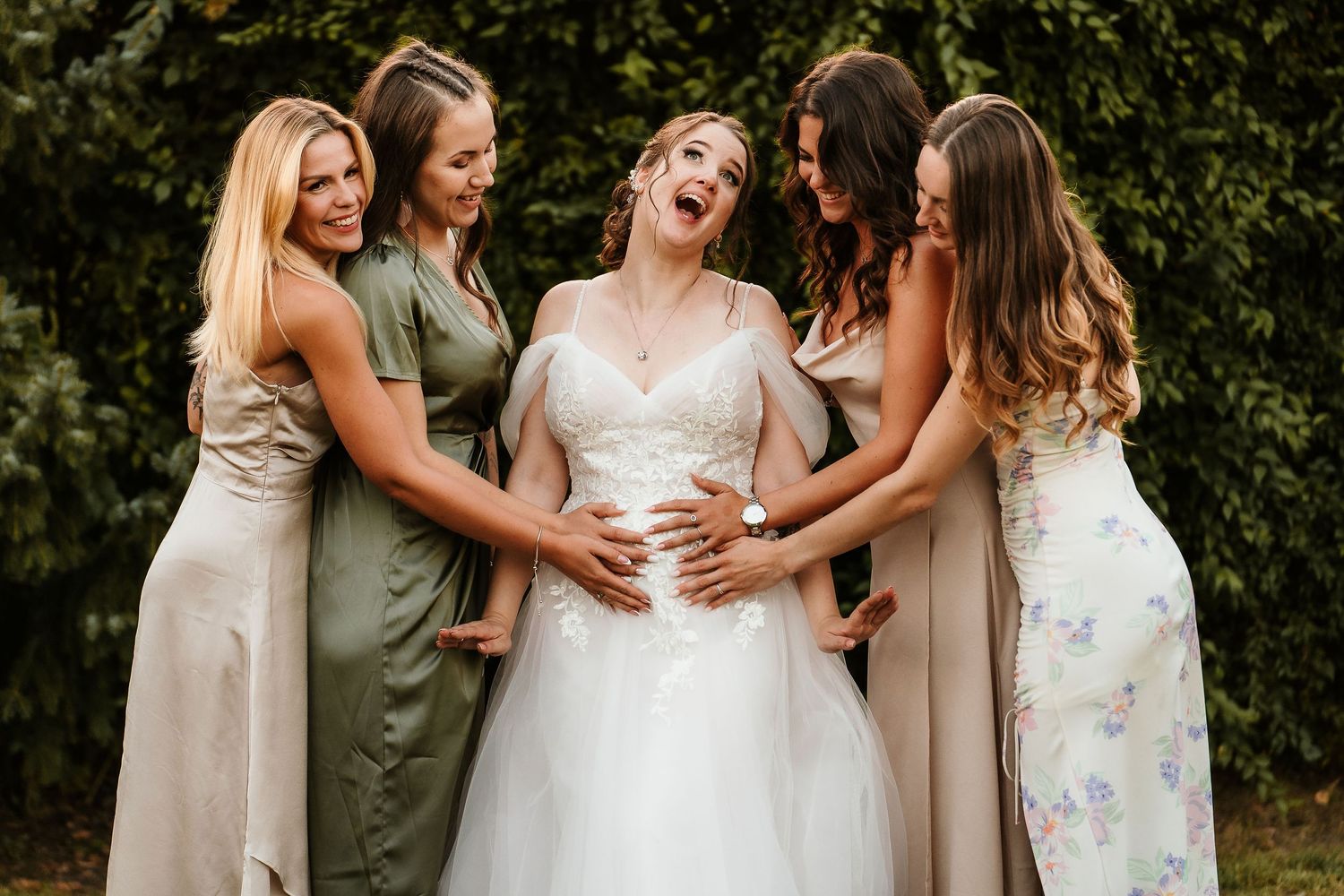 Bridal party sharing joyful moment while touching bride's belly outdoors.