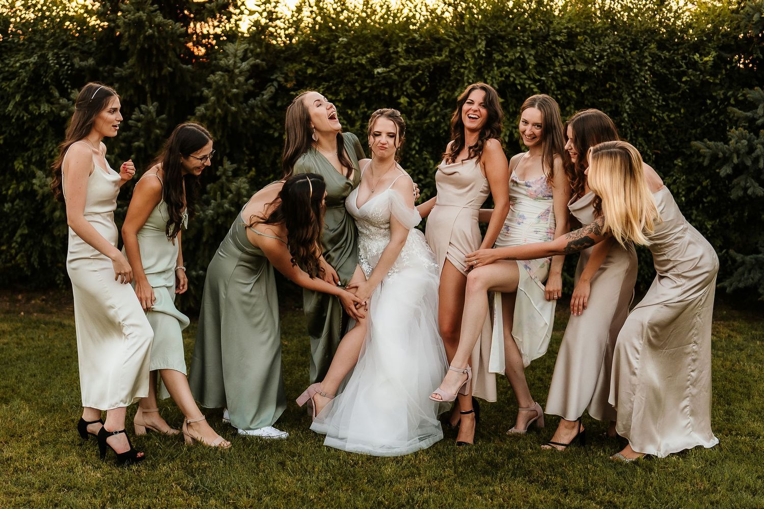 Wedding party in coordinated dresses laughing together on lawn at sunset.