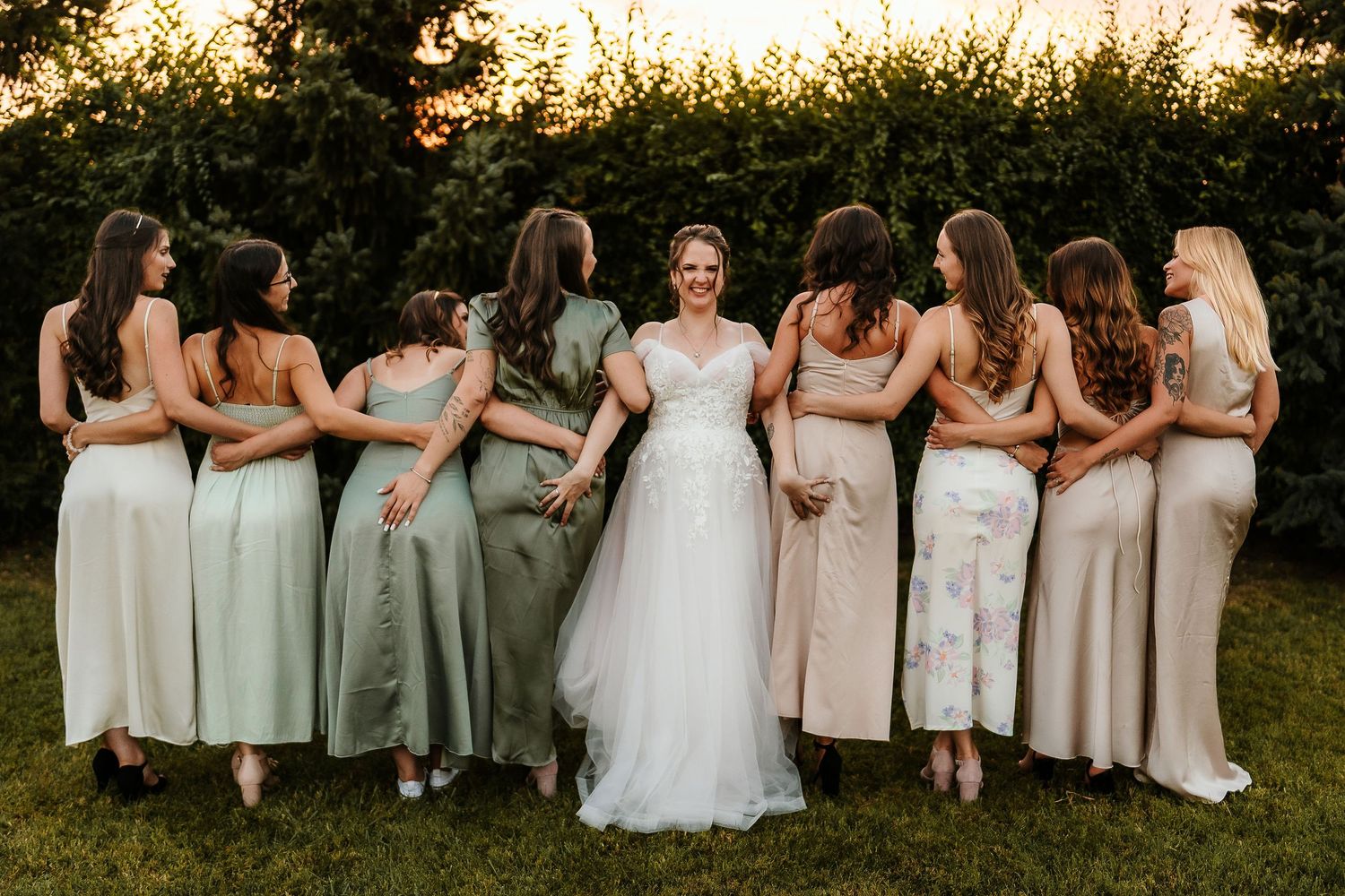 Bridal party in neutral and sage dresses posing with bride in garden setting.