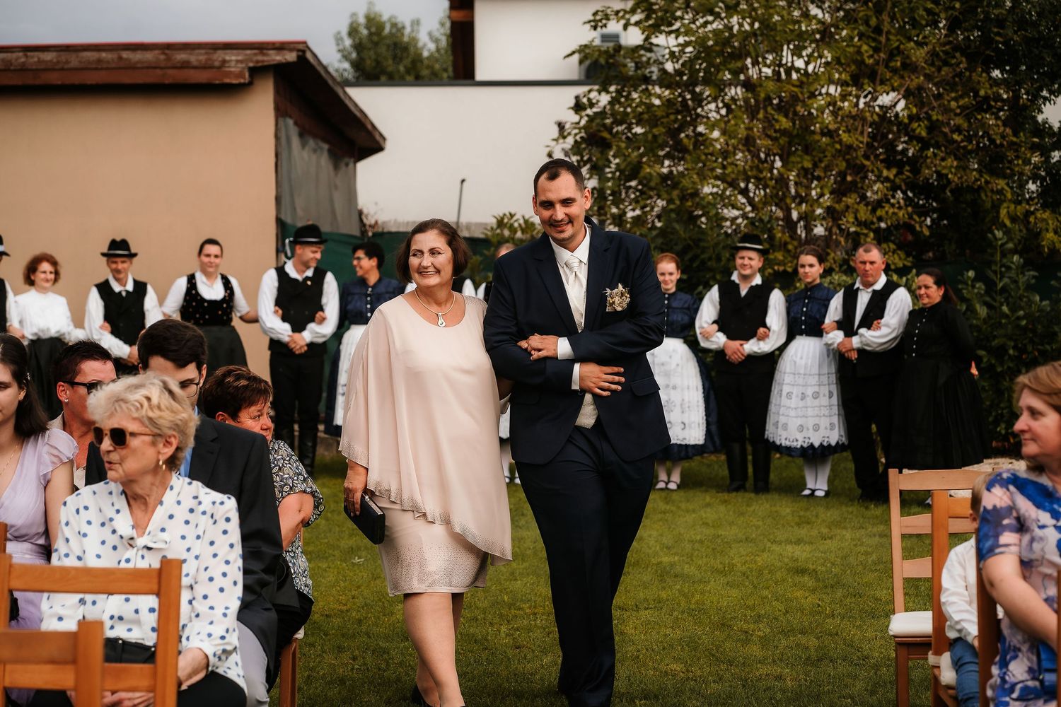 Wedding guests walking across a lawn with traditional folk performers in the background.