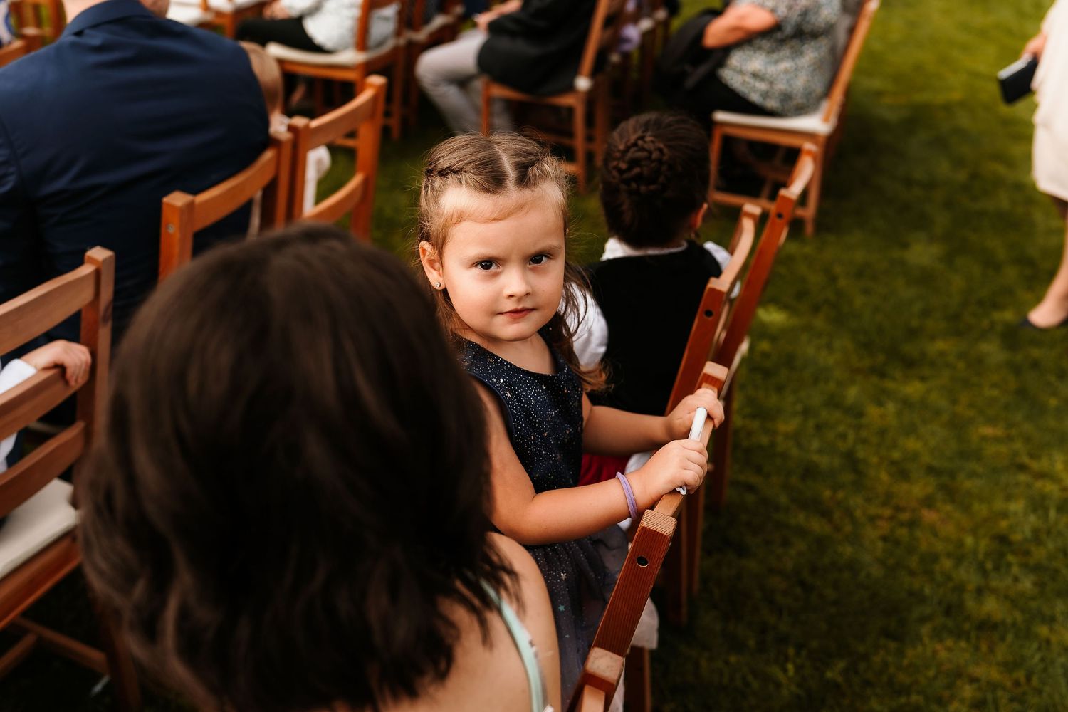 A young child sitting on wooden chairs during an outdoor wedding ceremony.