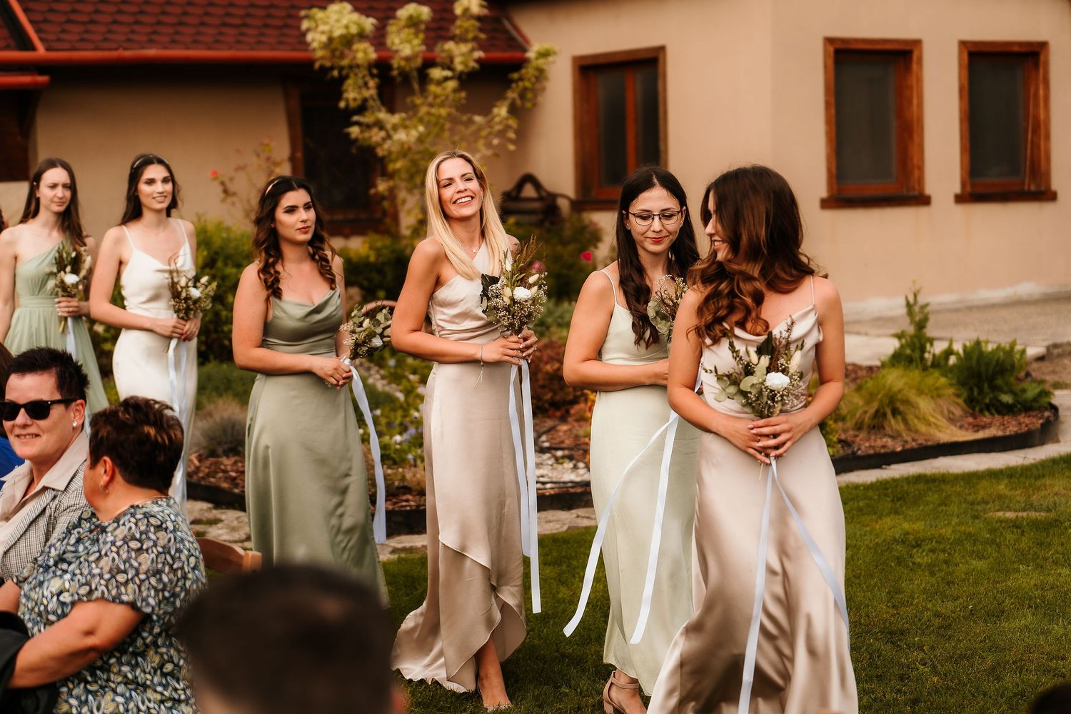Wedding party in coordinating sage and blush dresses gather on a sunny lawn.