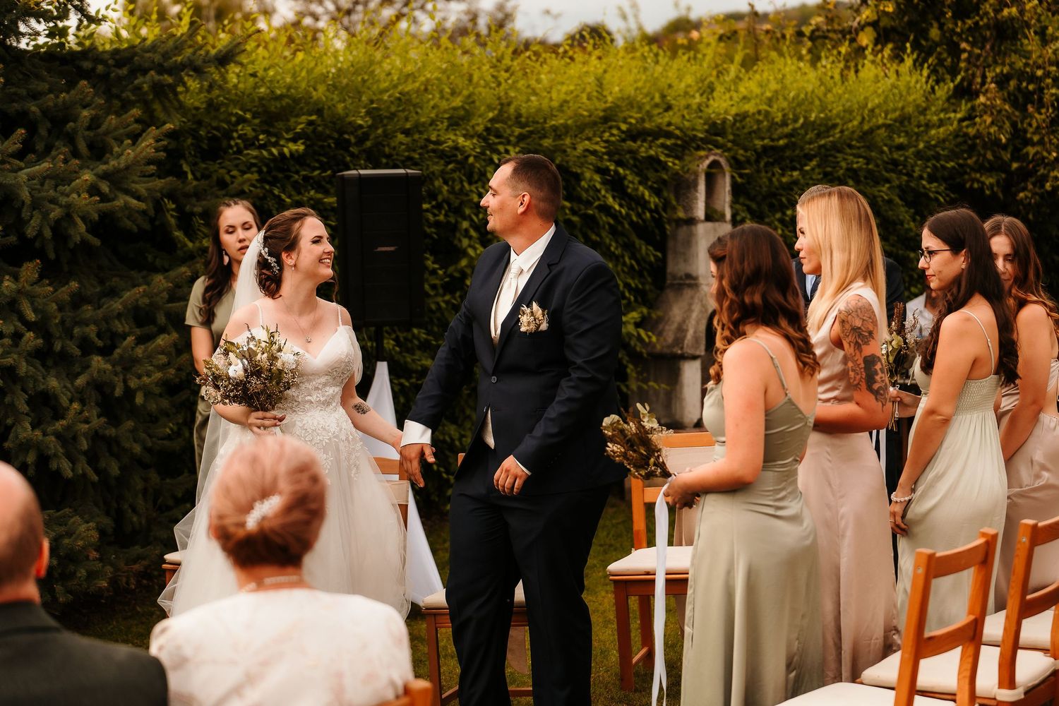 Outdoor wedding ceremony with couple and wedding party standing before garden backdrop.